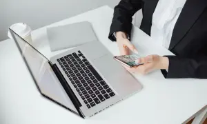 Kobieta w czarnej marynarce trzyma telefon, obok laptop i kubek. Zastanawia się, jak zaadresować pismo do adwokata.