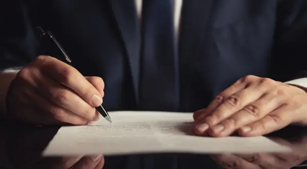 Mężczyzna w garniturze pisze długopisem na dokumencie. Dowiedz się, jak napisać testament bez notariusza.