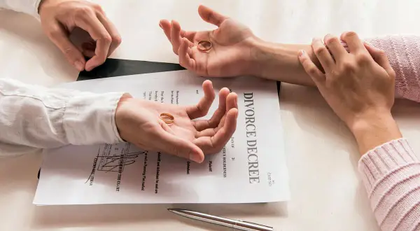 Dwie dłonie trzymają obrączki na tle dokumentu "DIVORCE DECREE". To symboliczny wniosek o rozwód za porozumieniem stron.