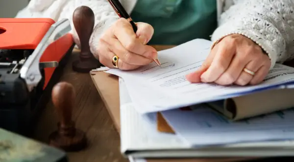 Ręce podpisują dokumenty, być może dotyczące spadku po matce bez testamentu. Obok leży stara maszyna do pisania i pieczątki.