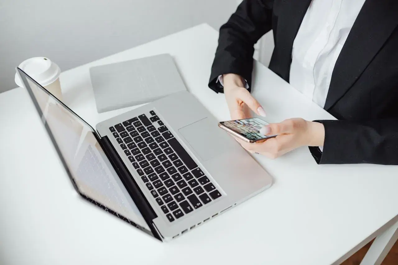 Kobieta w czarnej marynarce trzyma telefon, obok laptop i kubek. Zastanawia się, jak zaadresować pismo do adwokata.