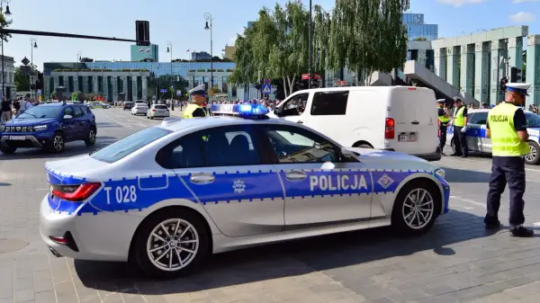 Radiowóz policyjny z włączonymi sygnałami świetlnymi na ulicy, obok policjanci.